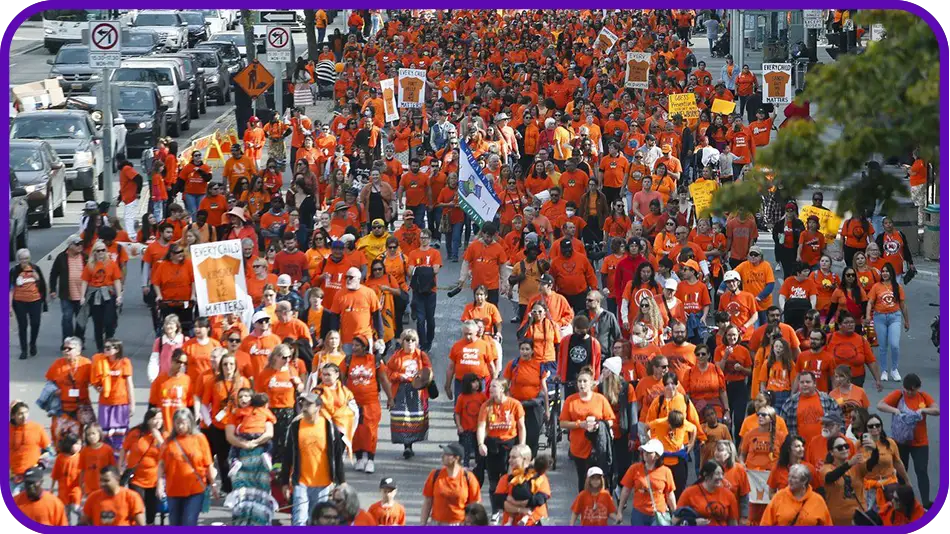 Large crowd of protesters wearing orange shirts marching down a city street with signs and banners.